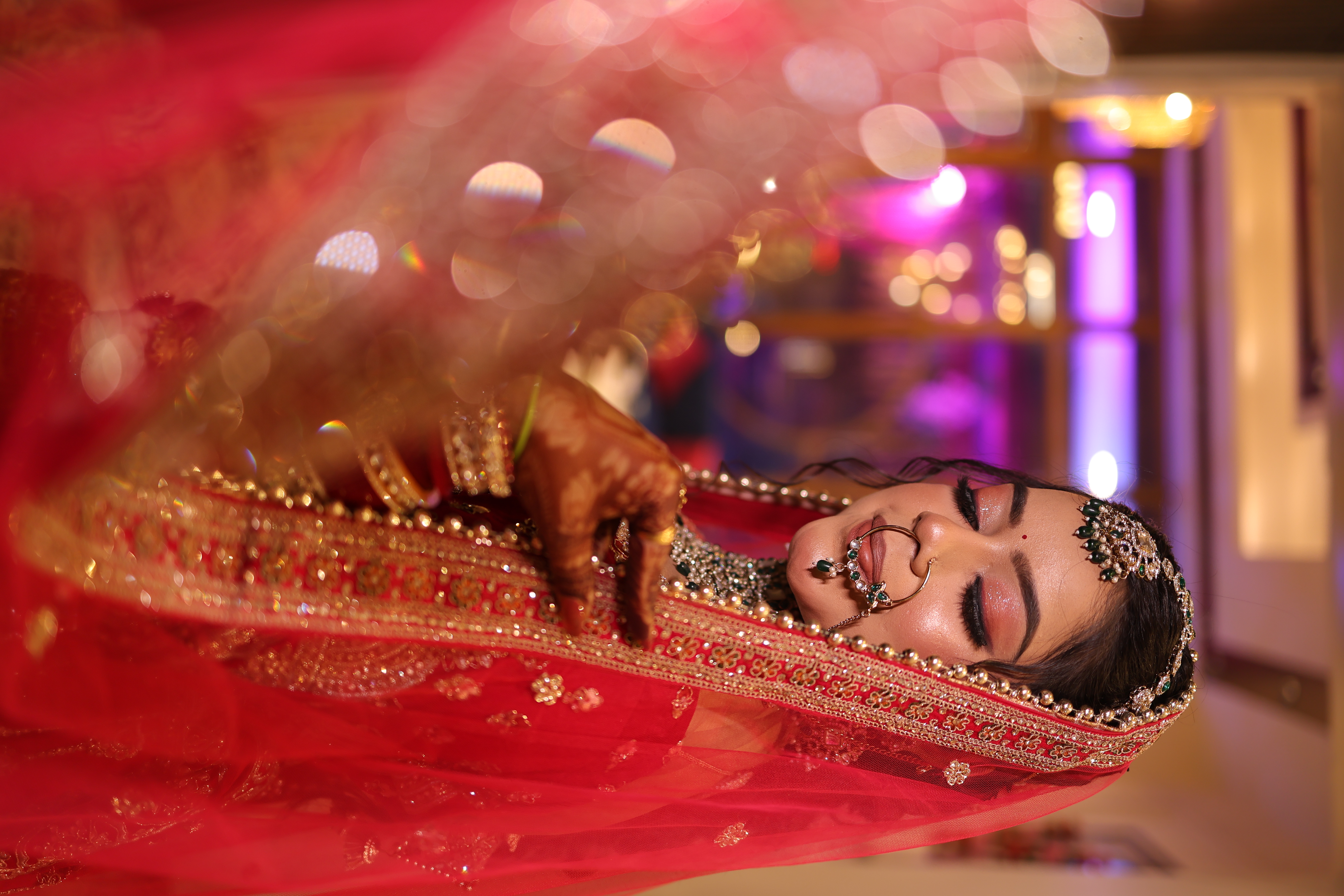 Ethereal bride with closed eyes showing glitter cut-crease eye glam and royal red lehenga with emerald jewelry